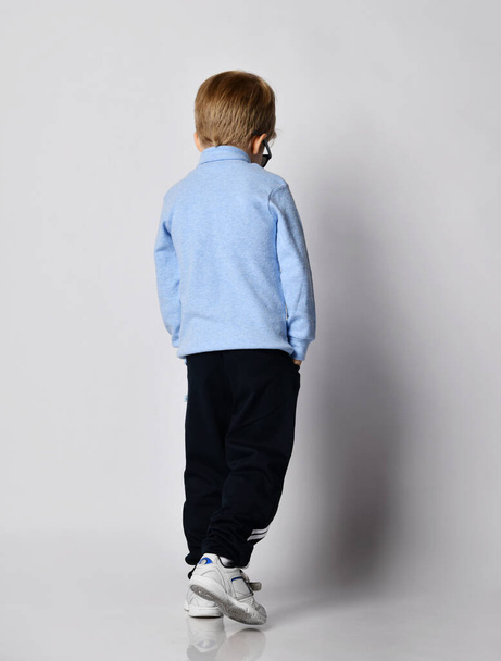 Blond kid boy in sneakers, black sport pants, blue turtleneck sweater stands back to us walks away looking down - Fotoğraf, Görsel