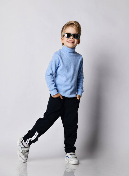Blond kid boy in sneakers, black sport pants, blue turtleneck sweater and sunglasses stands with his foot to the side - Photo, image