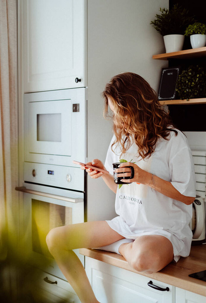 Young pretty woman drinks coffee sitting at her kitchen at home and using smartphone enjoys of morning. Smiling woman reading phone message - Фото, изображение