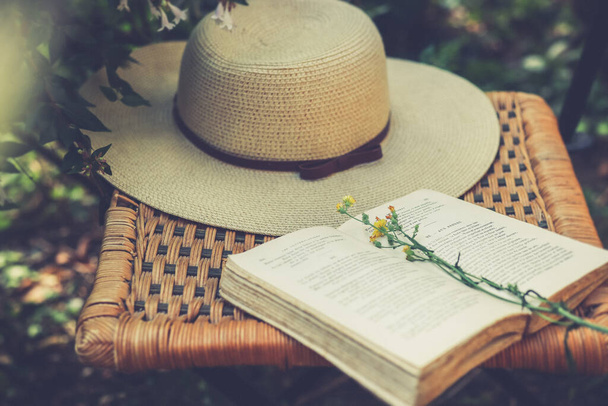 Полевые цветы на открытой книге и женская летняя шляпа на старом плетеном стуле в саду, ретро-фильтр, кашемировое ядро эстетики - Фото, изображение
