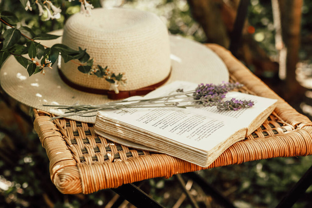 Лавандовый букет на открытой книге и женская летняя шляпа на старом плетеном стуле в саду, природе и медленной жизни концепции - Фото, изображение