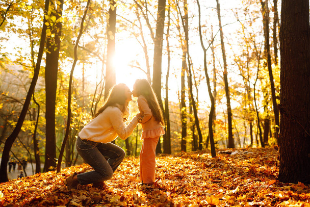 Stylish young Mother and daughter walking in the autumn forest at sunset. Mother and daughter embrace in autumn park. Mom and child having fun together in autumn. Family on a walk.  - Фото, изображение