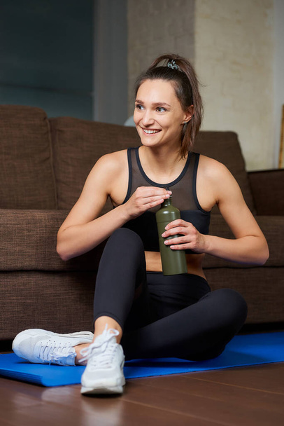 Uma menina desportiva em um terno apertado treino preto está sorrindo e abrindo uma garrafa de alumínio verde para saciar sua sede entre conjuntos de treinamento no tapete de ioga azul perto do sofá em casa. - Foto, Imagem
