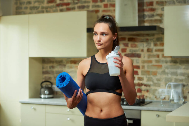 Ein lächelndes sportliches Mädchen im schwarzen Trainingsanzug hält die blaue Yogamatte und den weißen Plastikshaker in der Hand, um ihren Durst nach dem Training zu Hause zu stillen.. - Foto, Bild