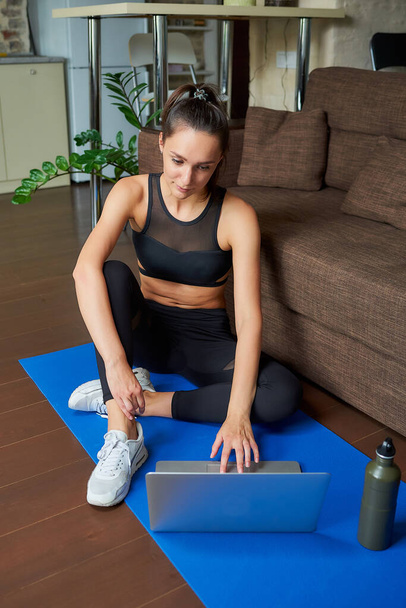 Uma menina esportiva em um terno apertado treino preto está trabalhando com um vídeo on-line em um laptop. Um treinador de fitness conduzindo uma aula de fitness virtual no tapete de ioga azul perto do sofá em casa
. - Foto, Imagem