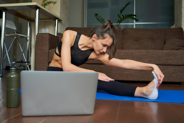 Antrenman kıyafetli sportif bir kız dizüstü bilgisayarında bir video ile esneme egzersizi yapıyor. Bir bayan fitness koçu evdeki kanepenin yanındaki mavi yoga minderinde uzaktan beden eğitimi dersi veriyor. - Fotoğraf, Görsel