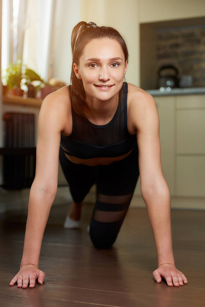 A smiling sporty girl in a black workout tight pants and top is relaxing after doing push-ups at home. A woman is practicing exercises for the chest at her apartment. - Photo, Image