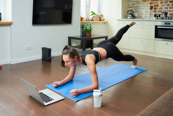 Una chica deportiva en un traje ajustado de entrenamiento está haciendo ejercicios en las nalgas con un video en línea en un ordenador portátil. Una entrenadora dirigiendo una clase de fitness remota en la alfombra azul de casa. - Foto, Imagen