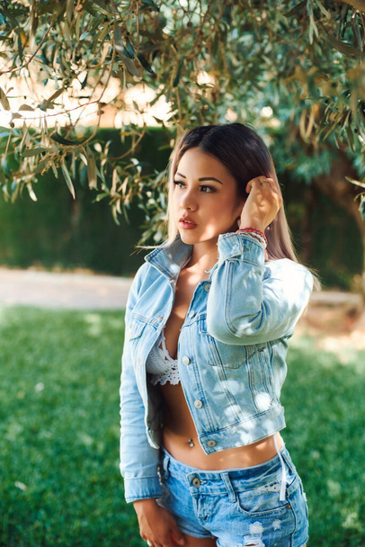 Beautiful blonde by the olive tree in a denim jacket and shorts in a light swimsuit. Beautiful portrait and light on the face - Фото, изображение