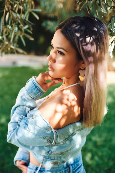 Beautiful blonde Korean woman by the olive tree in a denim jacket and shorts in a light swimsuit. Beautiful portrait and light on the face - 写真・画像