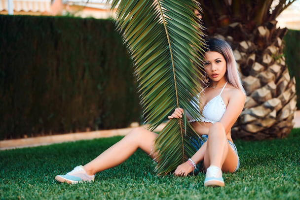 A beautiful blonde woman sits on the grass with a large palm branch in a white swimsuit. Beautiful portrait of a girl in a swimsuit - 写真・画像