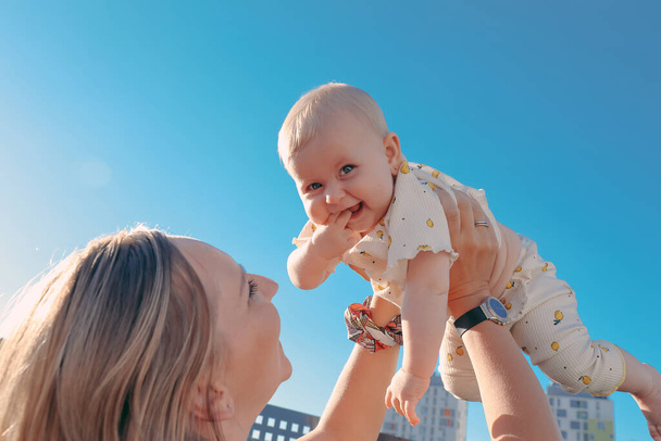 Portrait de mère soulevant son bébé à l'extérieur sous le ciel bleu. Fille heureuse souriant et regardant vers la caméra - Photo, image
