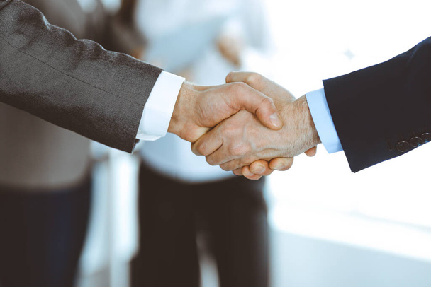 Business people shaking hands while standing with colleagues after meeting or negotiation, close-up. Group of unknown businessmen and women in modern office. Teamwork, partnership and handshake - Фото, изображение