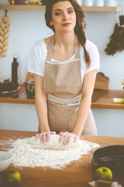 Young brunette woman cooking pizza or handmade pasta in the kitchen. Housewife preparing dough on wooden table. Dieting, food and health concept - Photo, image
