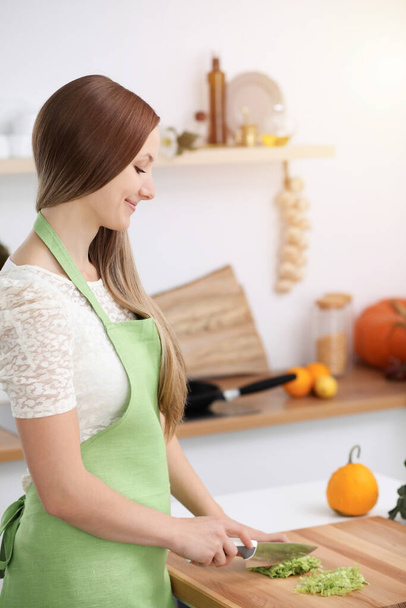 Beautiful woman dressed in apron is cooking meal in sunny kitchen - Foto, Imagem