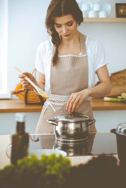 Young brunette woman cooking soup in kitchen. Housewife holding wooden spoon in her hand. Food and health concept - Valokuva, kuva
