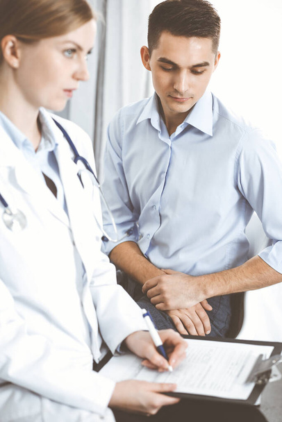Female doctor holding application form while consulting man patient in hospital office. Medicine and healthcare concept - Photo, image