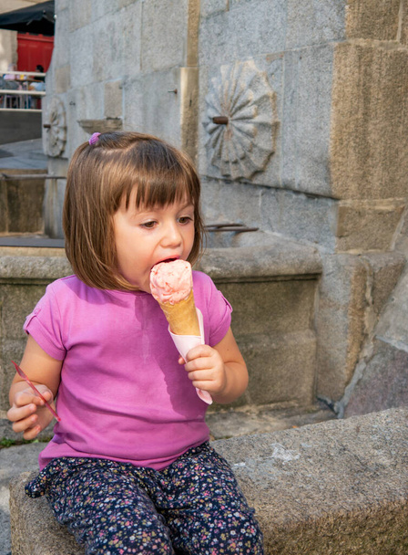 Pieni tyttö innokkaasti syö suuri mansikka jäätelöä istuu suihkulähde - Valokuva, kuva