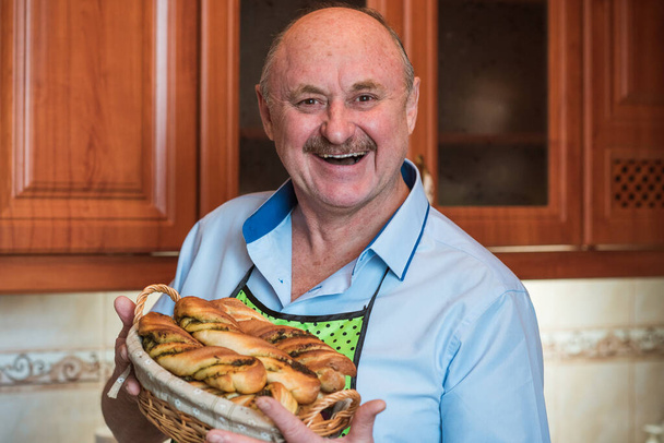 Happy smiling senior man on a kitchen with basket of bread, hobby and good mood in pensioner life - Photo, Image
