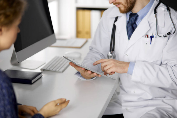 Unknown red-bearded doctor and patient woman discussing current health examination while sitting and using tablet computer in clinic, close-up. Medicine concept - Foto, Imagen
