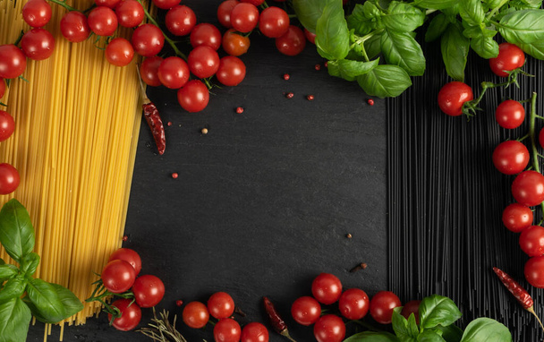 Raw black homemade spaghetti mockup with copy space on dark background. Dry black noodles, yellow macaroni bunch, cherry tomatoes, green basil. Uncooked italian spaghetto top view - Foto, imagen