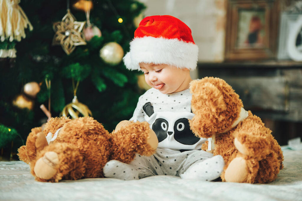 Pikkulapsi, jolla on joulupukin hattu sohvalla lelun kanssa. Ensimmäinen joulu. Uudenvuoden vapaapäivät. - Valokuva, kuva