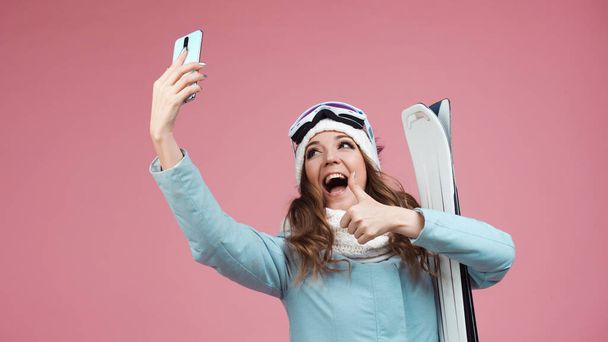 fun skier takes a selfie on her smartphone. A young woman with ski equipment in a jacket and hat, - Fotó, kép