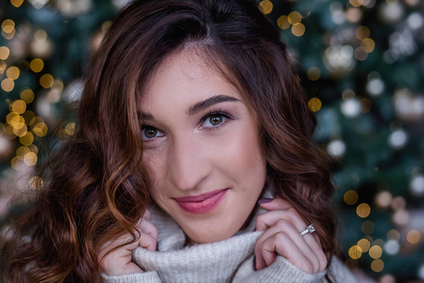 A beautiful curly-haired woman, wrapped in the collar of a gray sweater and smiles openly. A close-up portrait of a model looking at the camera. The background of a Christmas tree, boxes gifts, lights - Фото, изображение