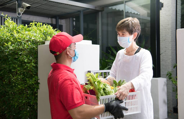 Азиатский курьер в маске и перчатках в красной форме доставляет коробку с продуктами питания, фруктами, овощами и напитками получателю во время вспышки КОВИД-19 - Фото, изображение