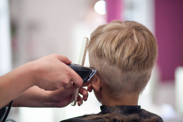 Um menino bonito senta-se em um cabeleireiro no estilista, uma criança em idade escolar está recebendo corte de cabelo em um salão de beleza, uma criança em uma barbearia, corte de cabelo de homens curtos. - Foto, Imagem
