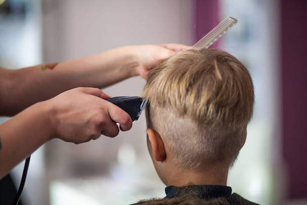 Mały uroczy chłopiec siedzi u fryzjera u stylisty, uczennica obcina włosy w salonie piękności, dziecko u fryzjera, krótka męska fryzura. - Zdjęcie, obraz