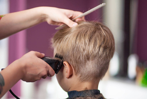 Маленький симпатичный мальчик сидит в парикмахерской у стилиста, школьник стрижется в салоне красоты, ребенок в парикмахерской, короткая мужская стрижка. - Фото, изображение