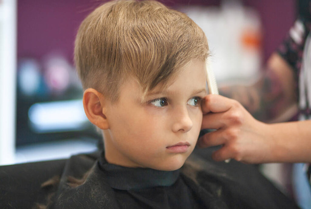 Маленький симпатичний хлопчик сидить у перукарні у стиліста, школярка стриже волосся в салоні краси, дитина в перукарні, коротка чоловіча зачіска
. - Фото, зображення