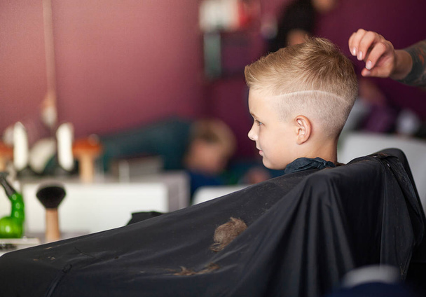 Un ragazzino carino si siede da un parrucchiere dallo stilista, uno scolaro si fa tagliare i capelli in un salone di bellezza, un bambino da un barbiere, un taglio di capelli corto da uomo. - Foto, immagini