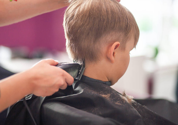 A little cute boy sits in a hairdresser's at the stylist's, a schoolchild is getting hair cut in a beauty salon, a child at a barbershop's, a short men's haircut. - Photo, Image
