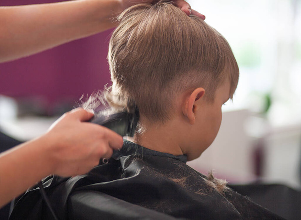 A little cute boy sits in a hairdresser's at the stylist's, a schoolchild is getting hair cut in a beauty salon, a child at a barbershop's, a short men's haircut. - Fotografie, Obrázek