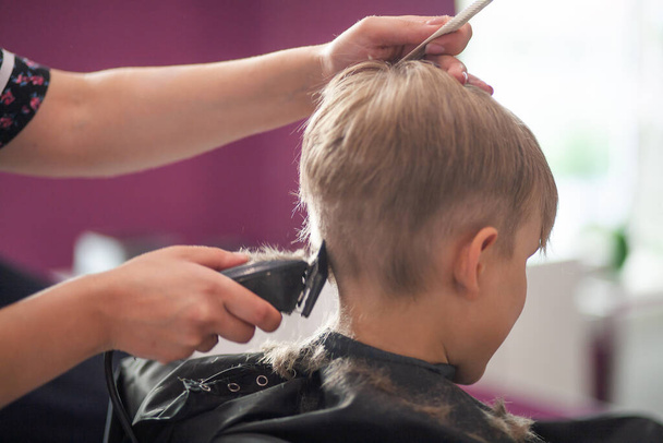 Un niño lindo se sienta en una peluquería en casa del estilista, un niño de la escuela se corta el pelo en un salón de belleza, un niño en una peluquería, un corte de pelo corto para hombres. - Foto, Imagen