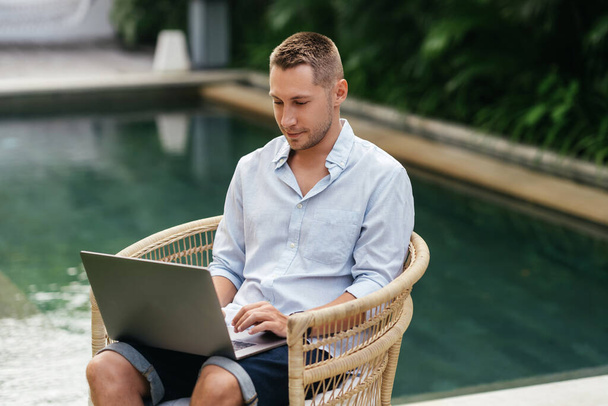 Young man using a laptop computer in a garden with a swimming pool. - Fotó, kép