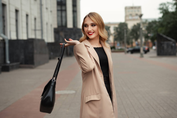 Porträt einer jungen schönen Frau mit langen blonden Haaren im beigen Trenchcoat und einer Tasche auf der Straße der Stadt. Modekonzept - Foto, Bild