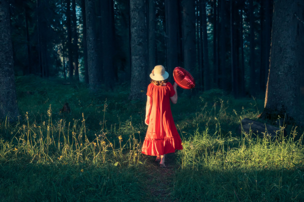 Ευτυχισμένη γυναίκα που απολαμβάνει το περπάτημα στην ειδυλλιακή φύση του δάσους, κρατώντας κόκκινο μπαλόνι σε σχήμα καρδιάς. Αγάπη και Ημέρα του Αγίου Βαλεντίνου έννοια. - Φωτογραφία, εικόνα