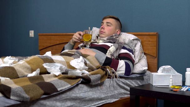 Ein junger Mann trinkt Tee mit Zitrone. Der Kerl liegt krank auf dem Bett und hat einen Schal um den Hals - Foto, Bild
