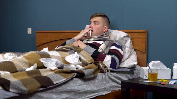 Молодий чоловік розбризкує ліки в носі. Хлопець хворий лежить на ліжку з шарфом на шиї
. - Фото, зображення