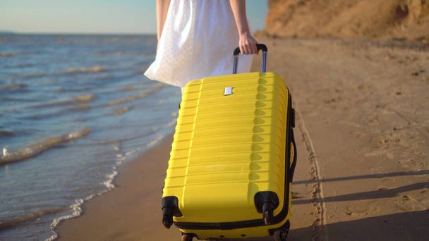 Νεαρή γυναίκα περπατά στην παραλία της θάλασσας με μια κίτρινη βαλίτσα κοντά. Ένα κορίτσι με λευκό φόρεμα περπατάει ξυπόλητο στην άμμο.. - Φωτογραφία, εικόνα