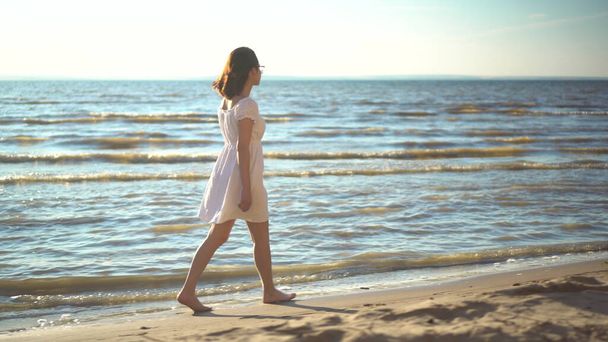 Attractive young woman walks along the sea beach. A girl in a white dress walks barefoot along the beach. - Photo, Image