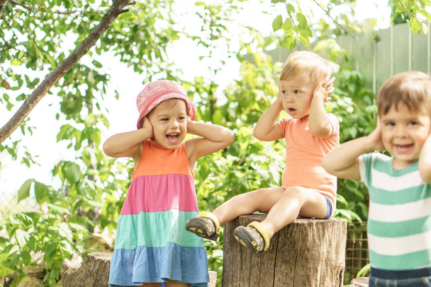 Дитинство, ігри, розвиток, сімейні концепції. Три дитини одного віку демонструють різні емоції. Щасливі усміхнені діти в саду грають на відкритому повітрі сестру і брати закривають вуха як глухі
 - Фото, зображення