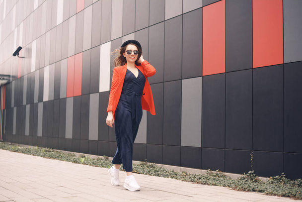 Девушка в осенней красной куртке идет на работу или в университет. Модель в комбинезоне на заднем плане здания. Осенняя мода - Фото, изображение