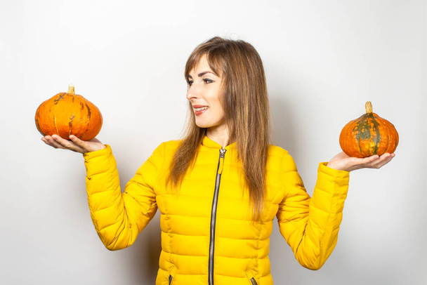 молодая девушка в желтой куртке, держащая две тыквы и глядя на тыкву на светлом фоне. Хэллоуин концепции, осень, празднование.  - Фото, изображение