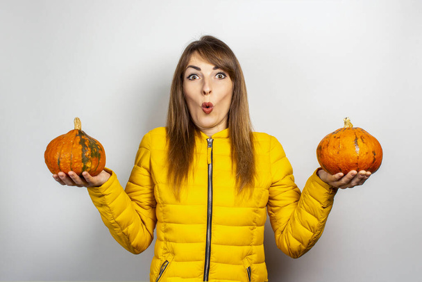 шокована молода дівчина в жовтій куртці тримає дві гарбузи на світлому фоні. концепція Хеллоуїна, осінь, святкування
. - Фото, зображення
