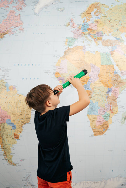 Biały chłopiec szkolny spoglądający w teleskop na mapę świata - Zdjęcie, obraz