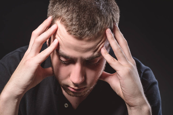 Young handsome man in a black shirt sits and holds his head on a black background. Experiencing stress, pain - 写真・画像